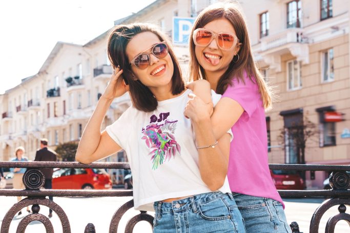 Zwei junge Frauen in DJF by Dean Jayson Freeman T-Shirts und Sonnenbrillen posieren fröhlich im Freien.
