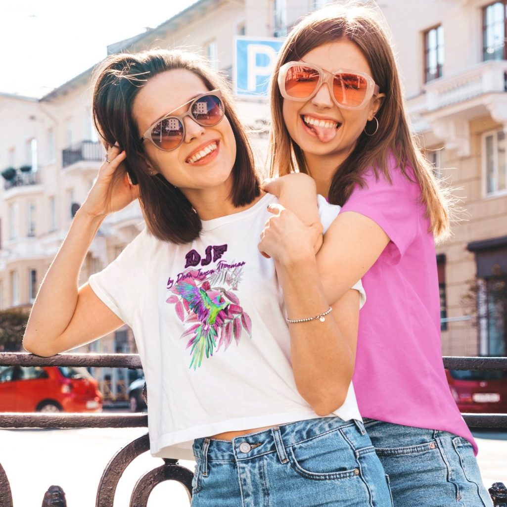 Zwei junge Frauen in Sonnenbrillen, lächelnd mit DJF by Dean Jayson Freeman T-Shirts mit Blick zur Kamera, vor einer urbana Kulisse.