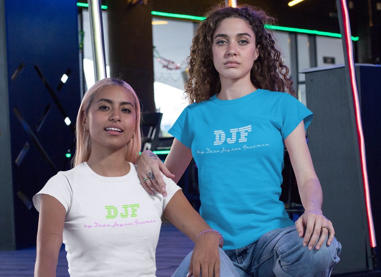 Zwei Frauen posieren in stylischen DJF by Dean Jayson Freeman T-Shirts vor einer bunten, beleuchteten Kulisse.