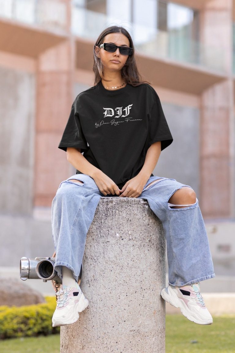 Frau sitzt auf einem Betonsäule, trägt ein schwarzes DJF by Dean Jayson Freeman T-Shirt und weite Jeans.